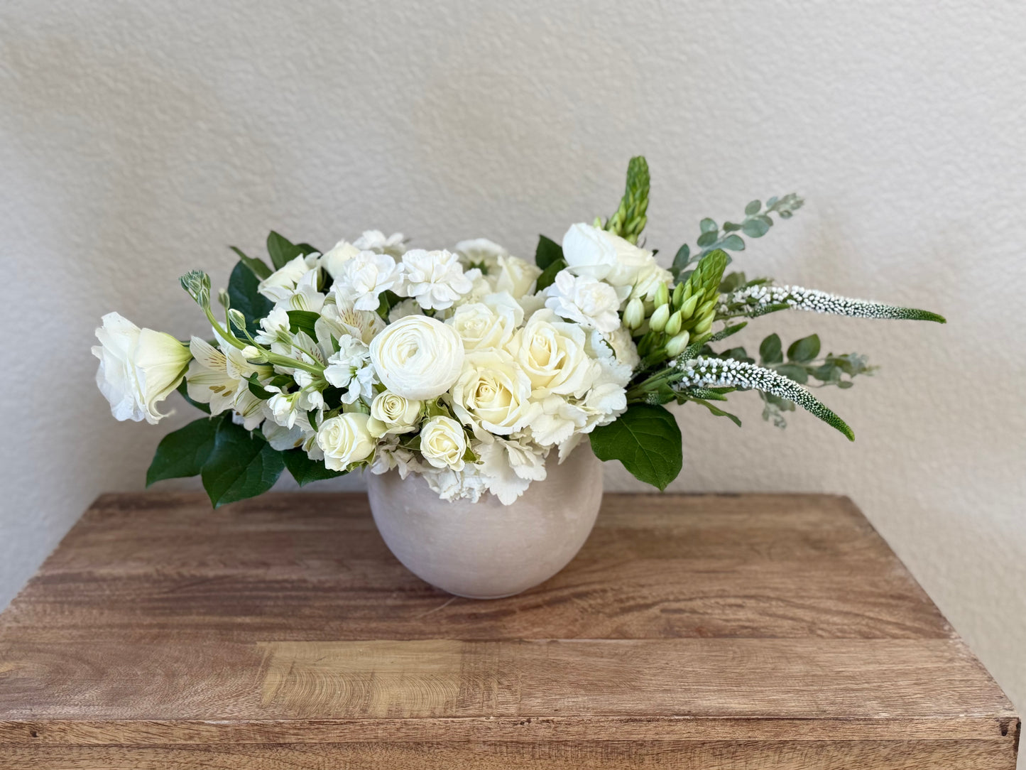 A floral arrangement of white roses, ranunculus, and green foliage in a round beige ceramic vase, displayed on a rustic wooden surface against a textured beige wall.